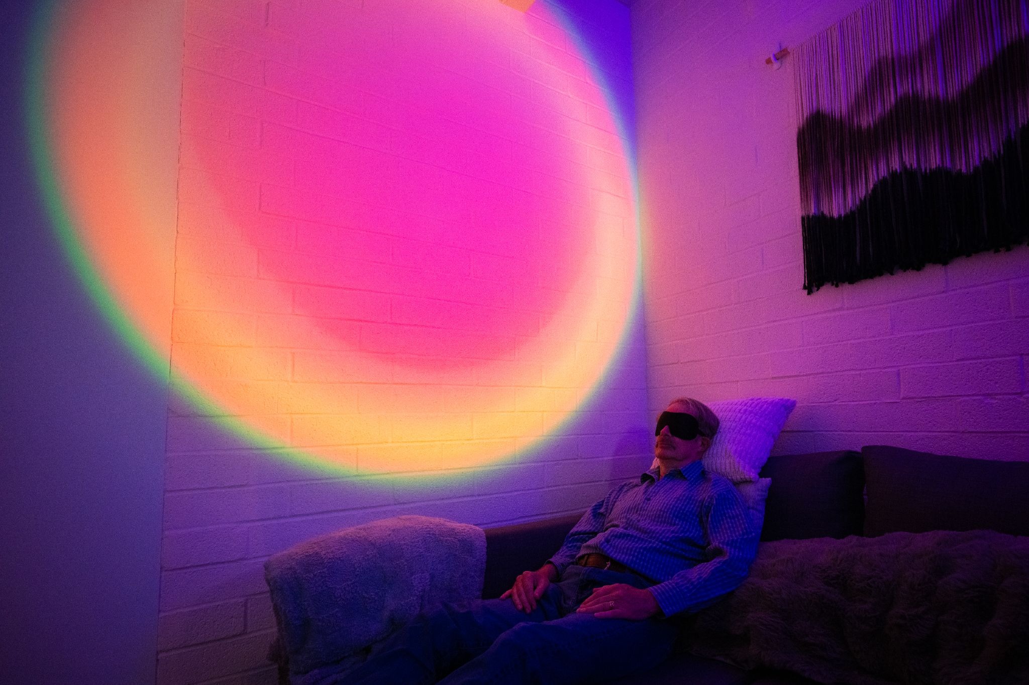 A man wearing an eye mask reclines on a couch in a darkened room. Behind him, the wall is illuminated with a circular, rainbow-colored light